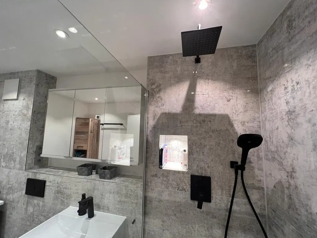 Fertiggestelltes Badezimmer mit grauen Großformatfliesen, schwarzen Armaturen und Regendusche