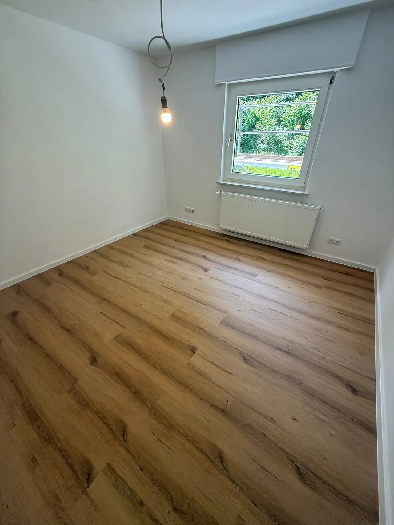 München, Zimmer nach der Kernsanierung: heller, leerer Raum mit Fenster, Heizkörper und Boden in Holzoptik
